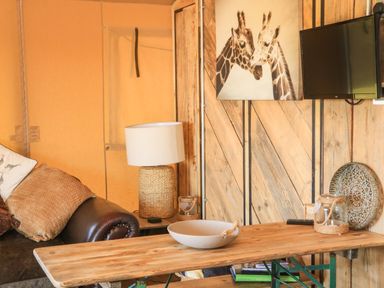 A room with a leather sofa and cushions a table lamp a wooden table wall art of giraffes a mounted television and decorative items at Semira Tent Lodge in Blackawton