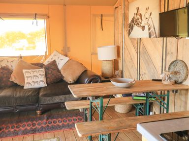A wooden interior with a brown sofa with animal print pillows a wooden table with benches a lamp and a wall mounted tv at Semira Tent Lodge in Blackawton