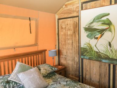 A bedroom with a wooden bed frame green patterned bedding a wooden nightstand with a gray lamp and a large bird painting on the wall at Semira Tent Lodge in Blackawton