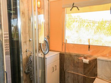 A bathroom with a glass shower enclosure a white sink cabinet a toilet and a window covered by a beige curtain at Semira Tent Lodge in Blackawton