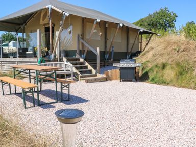 An outdoor area with a tent structure elevated on a wooden platform a picnic table with benches and a barbecue grill surrounded by gravel at Semira Tent Lodge in Blackawton