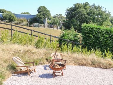 An outdoor area with two wooden chairs and a metal fire pit on a gravel surface with trees and fencing in the background at Semira Tent Lodge in Blackawton