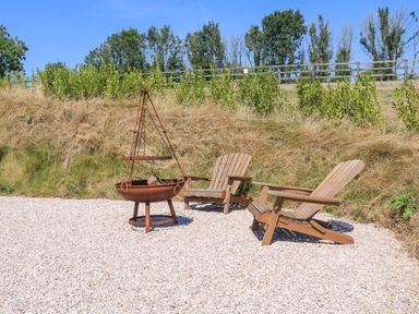 Two wooden chairs on gravel next to a metal fire pit with a grassy hill and trees in the background at Semira Tent Lodge in Blackawton