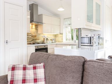 A kitchen with white cabinets and stone backsplash with a gray sofa and a red and white plaid pillow in the foreground at Honeysuckle Lodge in Thirlmere 14 White Cross Bay near Windermere