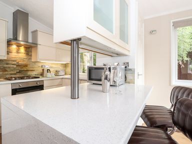 A kitchen with a white countertop island stools a microwave cocktail shaker and glass a stove and oven and stone backsplash at Honeysuckle Lodge Thirlmere 14 White Cross Bay near Windermere