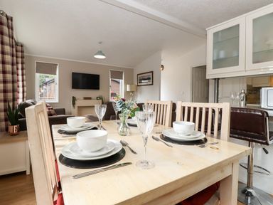 A dining table set with plates, bowls, cutlery, and glasses in a living room with a TV and windows at Honeysuckle Lodge, Thirlmere 14, White Cross Bay near Windermere