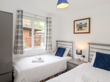 A bedroom with two single beds with blue pillows a nightstand with a lamp and a window with plaid curtains at Honeysuckle Lodge Thirlmere 14 White Cross Bay near Windermere