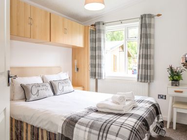 A bedroom with a bed, pillows, towel set, overhead cabinets, window with curtains, and a small vanity with flowers at Honeysuckle Lodge Thirlmere 14 White Cross Bay near Windermere