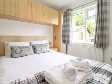 A bedroom with a bed with pillows and towels near a window with checkered curtains at Honeysuckle Lodge in Thirlmere 14 White Cross Bay near Windermere