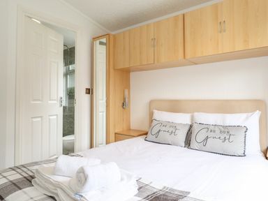 A bedroom with a bed with pillows saying be our guest and folded towels on it with wooden storage cabinets and a door to a bathroom at Honeysuckle Lodge Thirlmere 14 White Cross Bay near Windermere