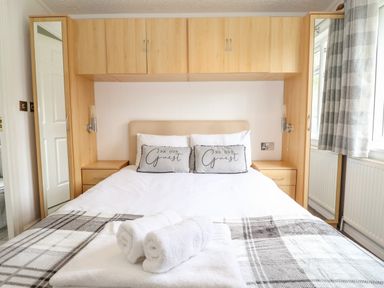 A bedroom with a bed with pillows and folded towels on it a wooden wardrobe with mirrors on each side and window with curtains at Honeysuckle Lodge Thirlmere 14 White Cross Bay near Windermere