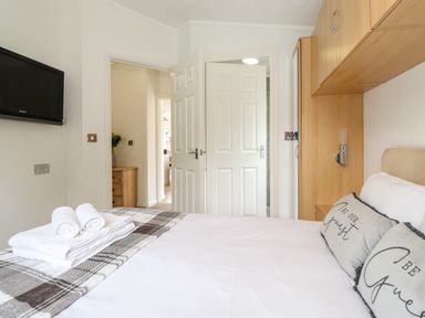 A bedroom with a double bed covered with white linens and plaid blanket with towels on top two doors open and a mounted tv on the wall at Honeysuckle Lodge Thirlmere 14 White Cross Bay near Windermere