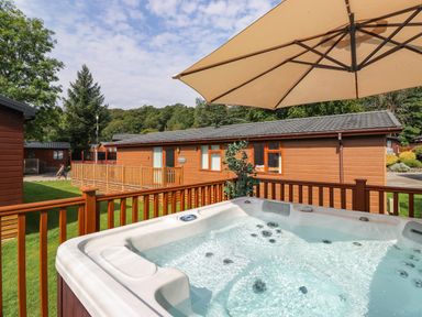 An outdoor hot tub on a wooden deck with an umbrella and a brown wooden cabin in the background at Honeysuckle Lodge in Thirlmere 14 White Cross Bay near Windermere