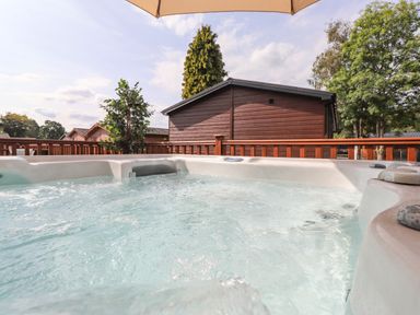 A hot tub on a wooden deck with trees and cabins in the background at Honeysuckle Lodge Thirlmere 14 White Cross Bay near Windermere