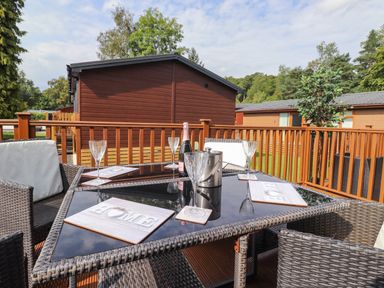 An outdoor patio with a glass table set with four glasses placemats and a bottle of champagne at Honeysuckle Lodge Thirlmere 14 White Cross Bay near Windermere