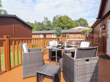 An outdoor wooden deck with wicker chairs around a glass table with a bottle and glasses at Honeysuckle Lodge Thirlmere 14 White Cross Bay near Windermere