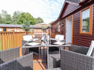 A wooden deck with a glass table and wicker chairs outside a wooden cabin at Honeysuckle Lodge Thirlmere 14 White Cross Bay near Windermere