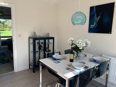 A dining room with a white table set for four with bowls glasses and cutlery a vase with white flowers a bottle of wine a hanging blue glass pendant light a glass cabinet with figurines and sculptures 2 Boathouse Terrace Mawgan Porth