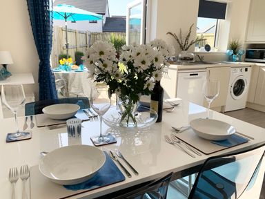 A dining table set with white bowls, wine glasses, cutlery, and a vase of white flowers in a kitchen with a washing machine and dishwasher at 2 Boathouse Terrace in Mawgan Porth