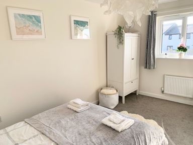 A bedroom with a bed with folded towels on a grey blanket a white wardrobe a basket a window with plants and grey curtains at 2 Boathouse Terrace in Mawgan Porth
