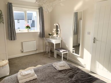 A bedroom with a bed, vanity table with lights, chair, full-length mirror, window with curtains, and a door at 2 Boathouse Terrace in Mawgan Porth