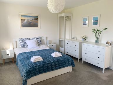 A bedroom with a white bed topped with blue blankets and pillows two white dressers an open wardrobe and framed pictures on the walls at 2 Boathouse Terrace in Mawgan Porth