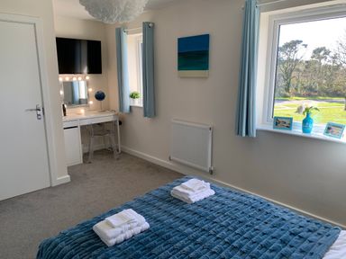 A bedroom with a bed covered with a blue blanket and white towels a desk with a mirror and chair a wall mounted TV and windows with blue curtains at 2 Boathouse Terrace in Mawgan Porth