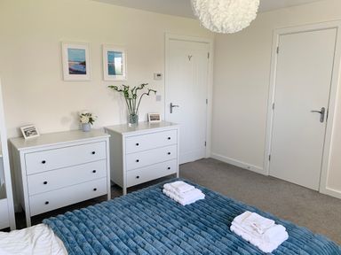 A bedroom with two white dressers plants framed pictures and towels on a blue bedspread at 2 Boathouse Terrace in Mawgan Porth