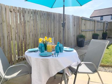 An outdoor patio with a round table covered with a white tablecloth blue plates and cups a pitcher of juice yellow flowers and two gray chairs under a blue umbrella at 2 Boathouse Terrace in Mawgan Porth
