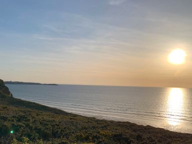 A coastal landscape with the sun setting over the ocean and vegetation in the foreground at 2 Boathouse Terrace in Mawgan Porth