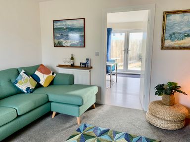 A living room with a green sofa and colorful cushions two paintings on walls and a round wicker side table with a plant at 2 Boathouse Terrace Mawgan Porth