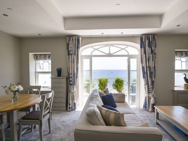 A living room with a beige sofa blue pillows wooden dining table chairs and a view of the sea through large windows at 1 Argyle House Lyme Regis
