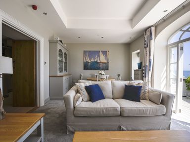A living room with a sofa and blue pillows a dining table with chairs and a painting of sailboats on the wall at 1 Argyle House in Lyme Regis