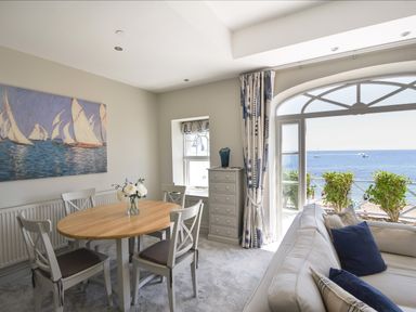 A dining area with a wooden table and chairs next to a living room with a sofa and a large window overlooking the sea at 1 Argyle House in Lyme Regis