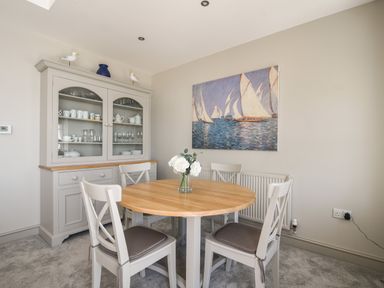 A dining area with a round wooden table and four chairs a cabinet with dishes and glassware and a painting of sailboats on the wall at 1 Argyle House in Lyme Regis
