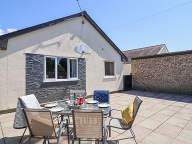 An outdoor patio area with a glass dining table and six chairs near a house with two windows and a basketball hoop at Ty Glan Morfa in Morfa Bychan