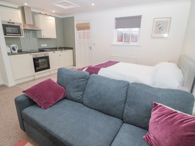 A small studio with a grey sofa with purple pillows a bed with white and purple bedding a kitchen area with white cabinets and appliances and a window with a grey blind at Cherry Studio in Crookham near Cornhill On Tweed