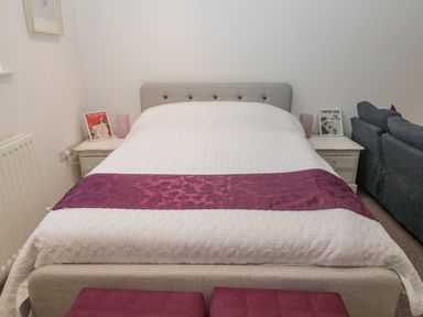 A bedroom with a bed with white quilt and purple runner and bedside tables with framed pictures at Cherry Studio in Crookham near Cornhill-On-Tweed