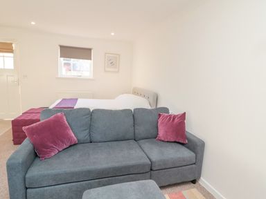 A room with a gray sofa with pink pillows a bed with white bedding and a purple throw and a window at Cherry Studio in Crookham near Cornhill-On-Tweed