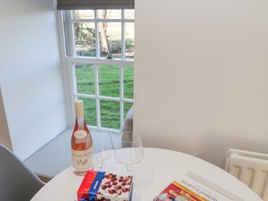 A table with a bottle of wine, two wine glasses, a box of chocolates and a welcome guide next to a window with a seat at Cherry Studio in Crookham near Cornhill-On-Tweed