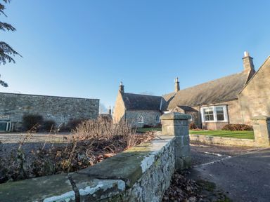A stone house with chimneys and a garden behind a low stone wall at Cherry Studio in Crookham near Cornhill-On-Tweed
