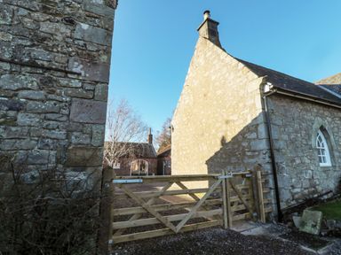 A wooden gate between stone buildings with a tree and brick building in the background at Cherry Studio in Crookham near Cornhill On Tweed