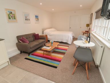 A room with a brown sofa with purple pillows a bed with white bedding a wooden coffee table on a colorful striped rug a small round white dining table with two gray chairs and a window with blinds at Silverbirch in Crookham near Cornhill-On-Tweed