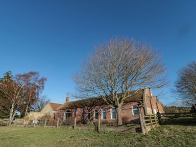 A brick and stone building with a tiled roof surrounded by a fence and trees in Crookham near Cornhill-On-Tweed