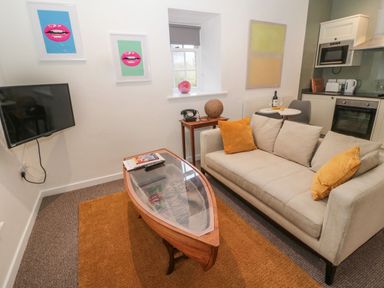 A living room with a beige sofa with orange pillows a boat shaped glass coffee table and a wall mounted TV at Cheviot View in Crookham near Cornhill-On-Tweed