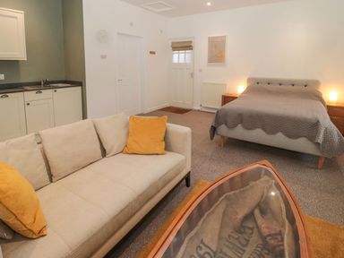 An open plan room with a beige sofa with yellow cushions a bed with lamps on wooden bedside tables a kitchenette with a sink and cupboards and a wooden coffee table with a glass top at Cheviot View in Crookham near Cornhill-On-Tweed