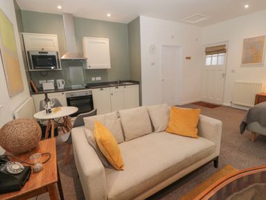 An open plan living space with a beige sofa with yellow cushions a small dining table with chairs and a kitchenette with white cabinets and appliances at Cheviot View in Crookham near Cornhill-On-Tweed