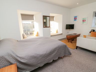 A bedroom with a bed covered with a gray quilt near two window seats and a small sitting area with a sofa and a wooden coffee table at Cheviot View in Crookham near Cornhill-On-Tweed