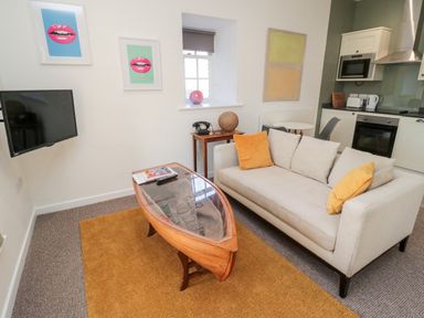 A living room with a beige sofa and yellow cushions a wooden boat-shaped coffee table and a small kitchen area at Cheviot View in Crookham near Cornhill-On-Tweed