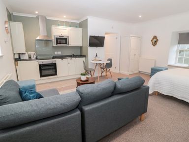 A studio apartment with a kitchen area a grey sofa a round dining table with two grey chairs a white wardrobe and a bed with a white cover at Flodden in Crookham near Cornhill-On-Tweed